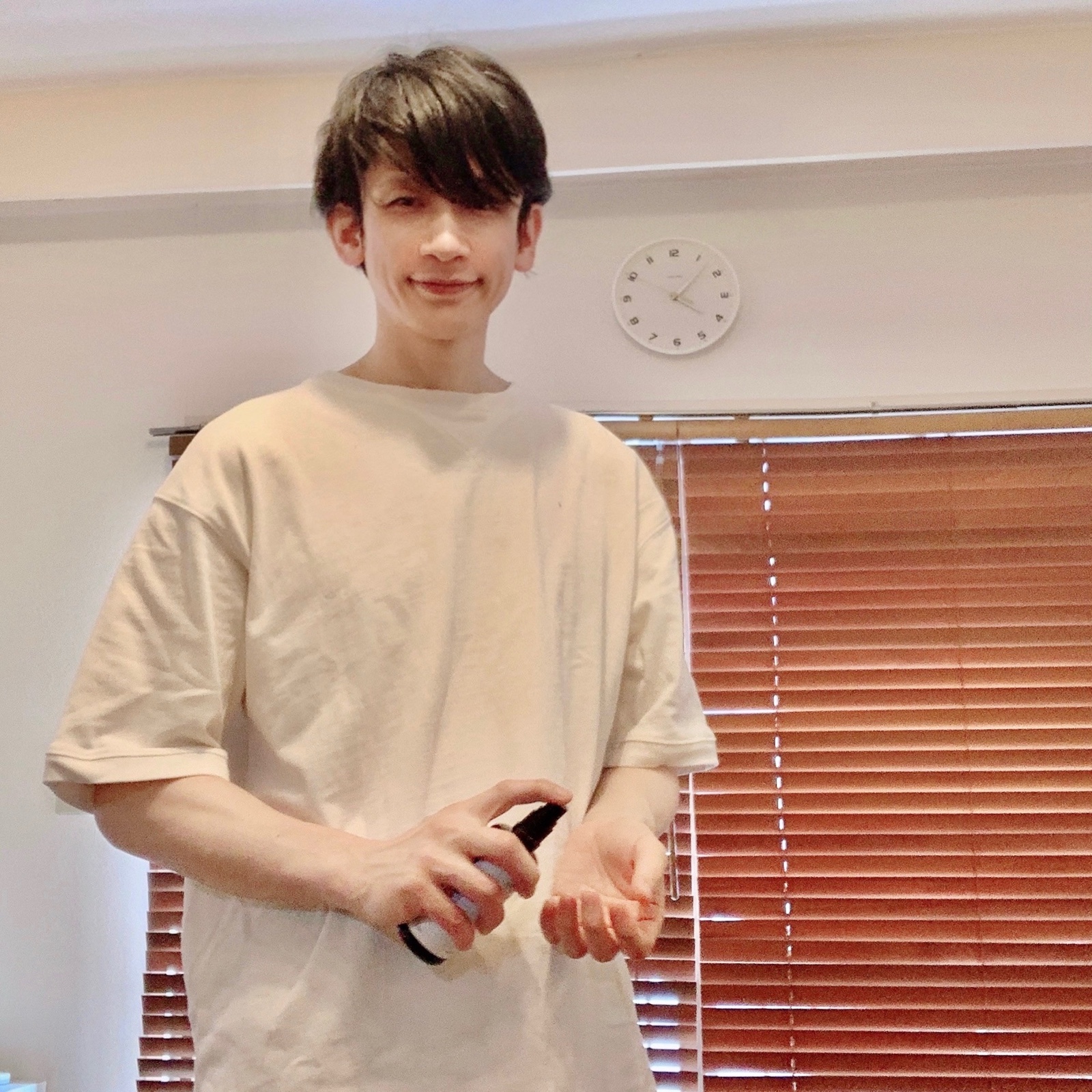 ムーンテッド(MoonTed)の日記より、スプレーボトルを手に立つムーンテッド(MoonTed)のセラピストAKI。ベージュのTシャツ姿、奥に時計とブラインド