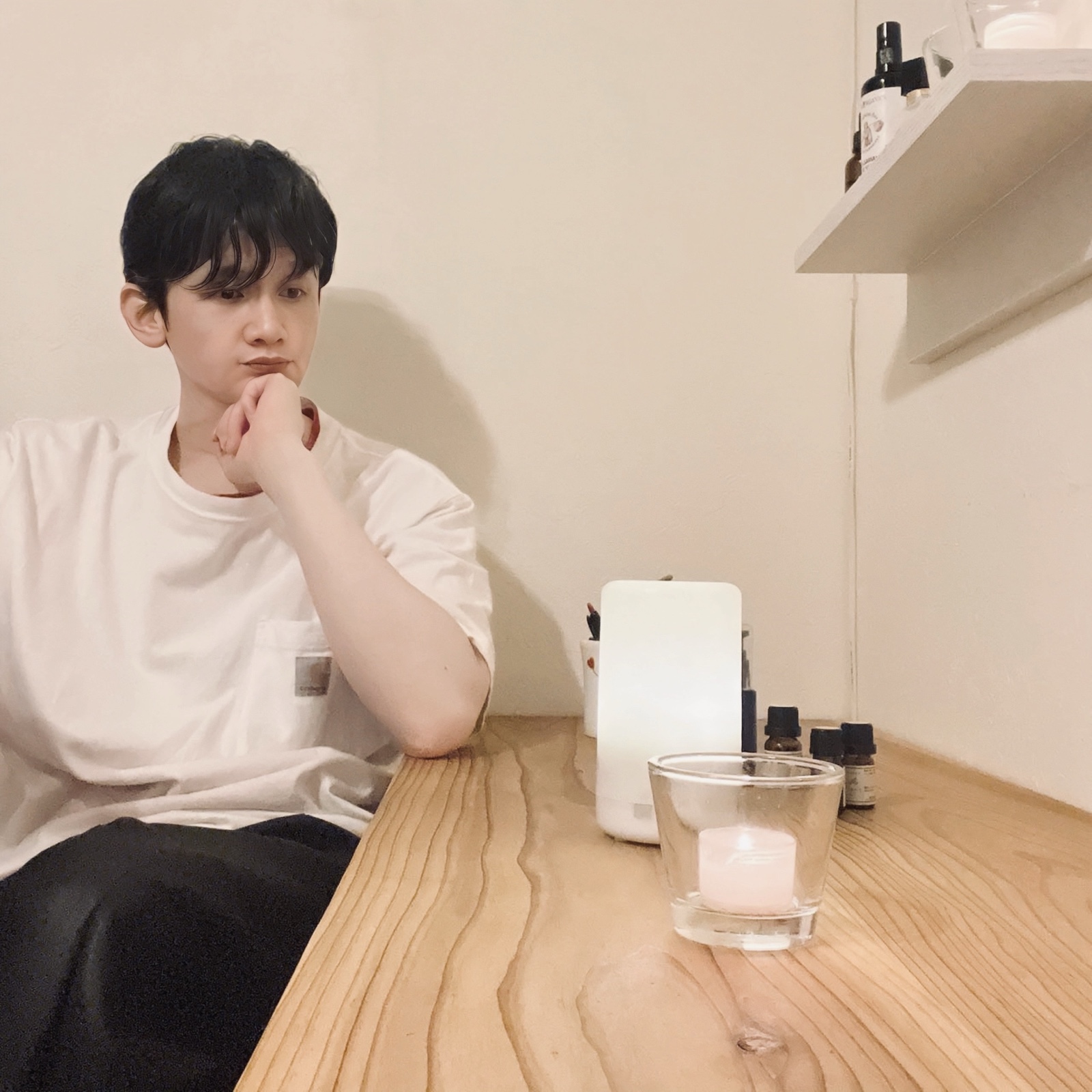 ムーンテッド(MoonTed)の日記より、机に頬杖をついて考え込むムーンテッド(MoonTed)のセラピストAKI。手前にキャンドル、棚に遮光瓶