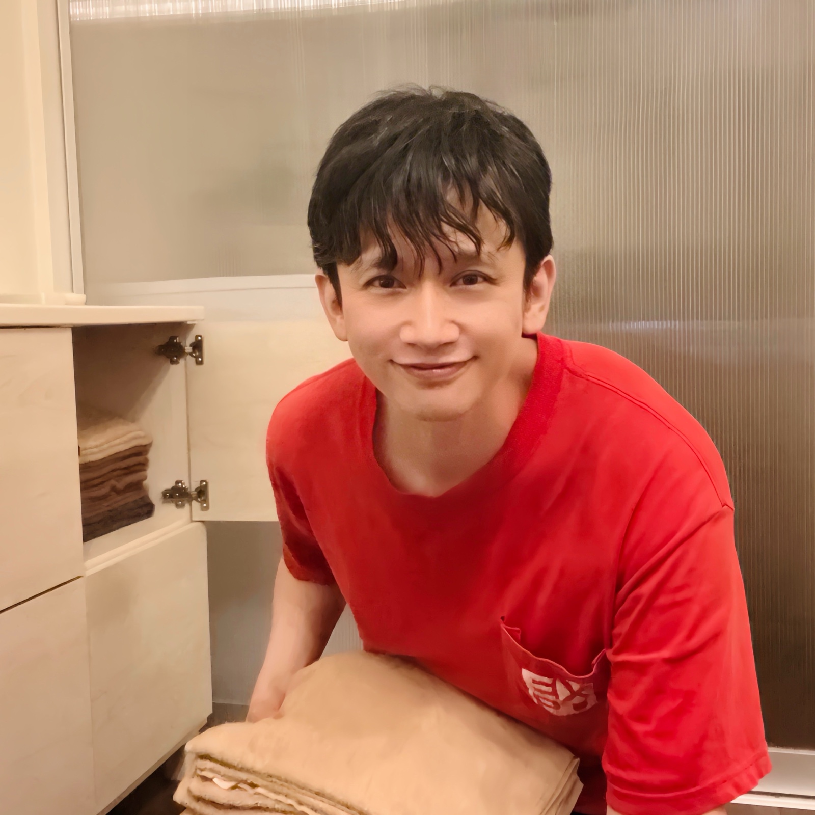 ムーンテッド(MoonTed)の日記より、たたんだ毛布を抱えて笑うムーンテッド(MoonTed)のセラピストAKI。赤いTシャツ姿、横の棚にタオル