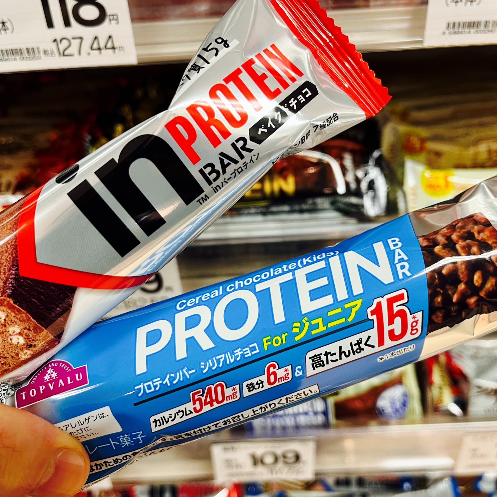 ムーンテッド(MoonTed)の日記より、スーパーの棚の前で手に持った2本のプロテインバー。森永のinバーと、トップバリューのプロテインバー