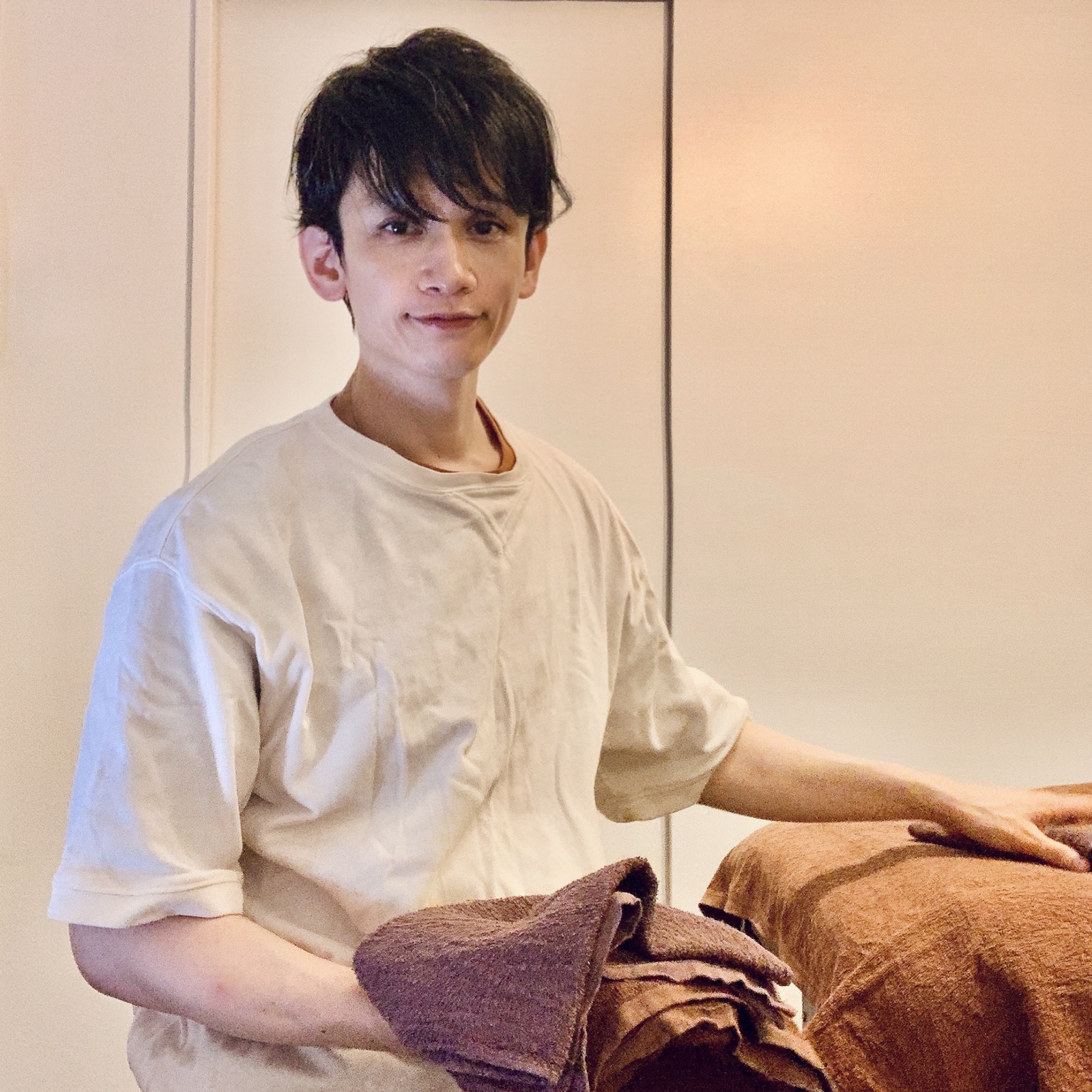 ムーンテッド(MoonTed)の日記より、タオルを抱え、施術台に手を置いて微笑むムーンテッド(MoonTed)のセラピストAKI。ベージュのTシャツ姿