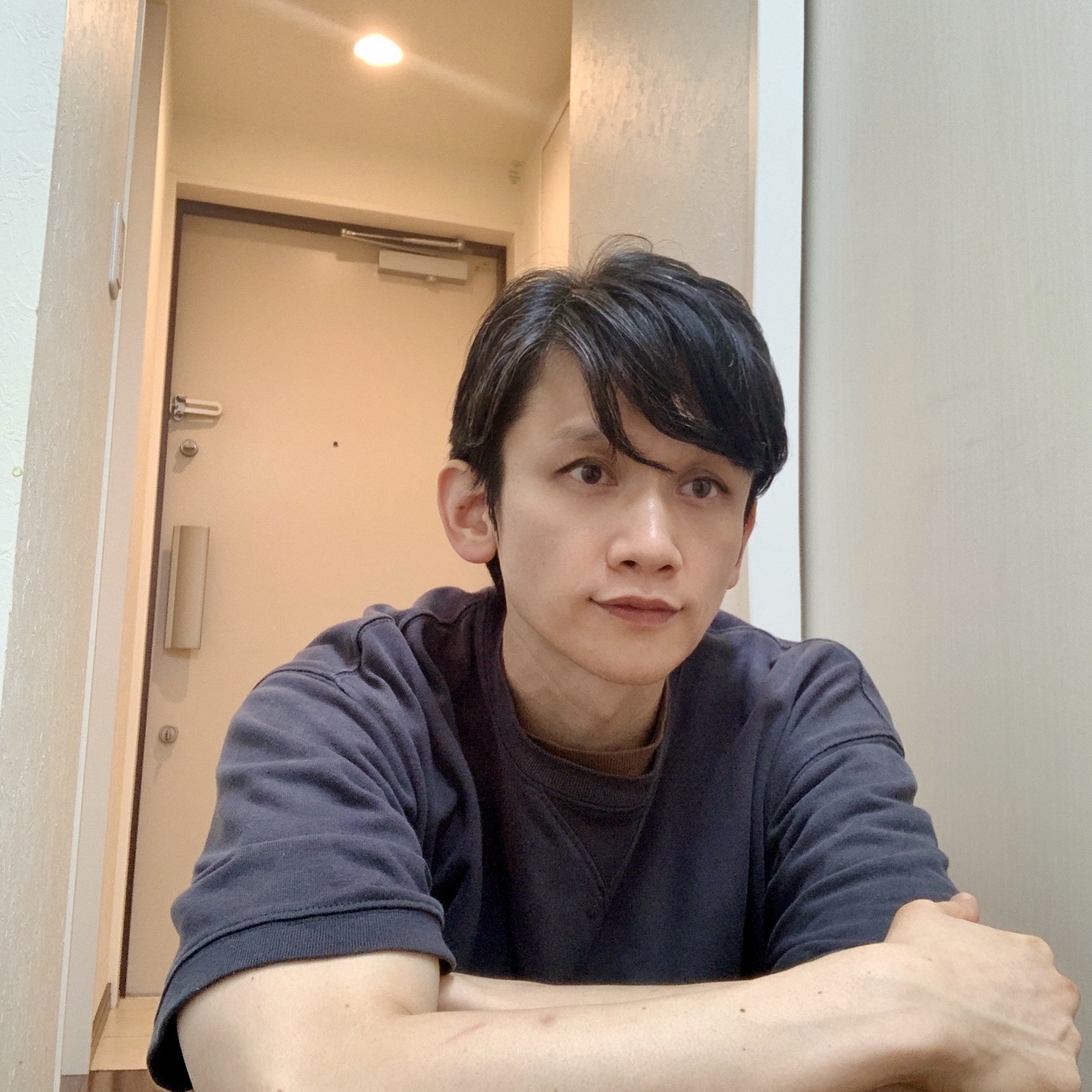 ムーンテッド(MoonTed)の日記より、腕を組んでテーブルにもたれるムーンテッド(MoonTed)のセラピストAKI。紺のTシャツ姿、奥に玄関のドア