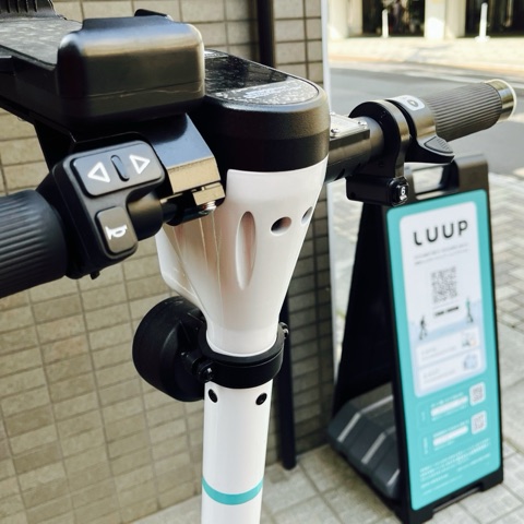 ムーンテッド（MoonTed）の日記より、LUUPの電動キックボードのハンドル。手元にボタン式のアクセル、奥にLUUPのポートの看板