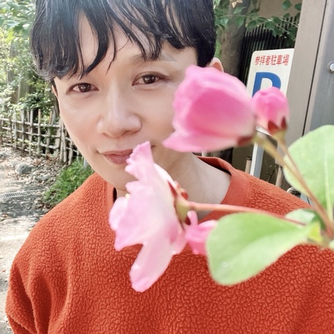 ムーンテッド（MoonTed）の日記より、代々木八幡宮で、ピンクの花越しに微笑むムーンテッド（MoonTed）のセラピストAKI。オレンジのセーター姿