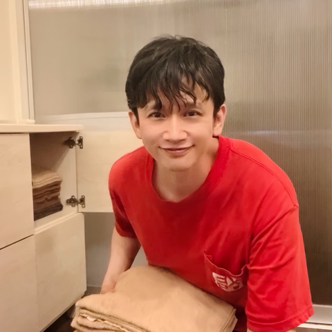 ムーンテッド（MoonTed）の日記より、たたんだ毛布を抱えて笑うムーンテッド（MoonTed）のセラピストAKI。赤いTシャツ姿、横の棚にタオル
