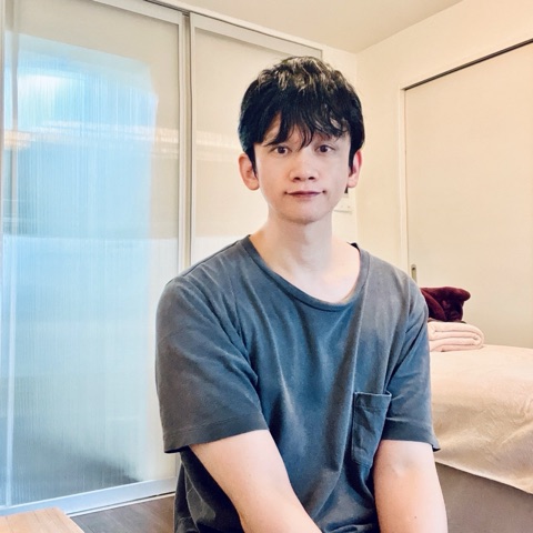 ムーンテッド（MoonTed）の日記より、施術室で、施術台の横に座って微笑むムーンテッド（MoonTed）のセラピストAKI。グレーのTシャツ姿