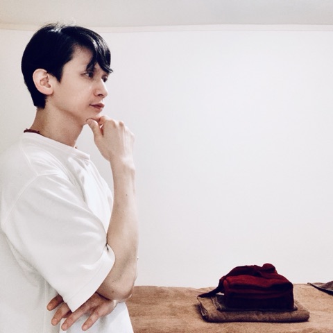 ムーンテッド（MoonTed）の日記より、施術台の横で、あごに手をあてて考えるムーンテッド（MoonTed）のセラピストAKI。白いTシャツ姿、施術台に赤いブランケット
