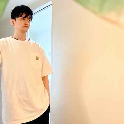 ムーンテッド（MoonTed）の日記より、窓辺に立ち、横を見るムーンテッド（MoonTed）のセラピストAKI。白いTシャツ姿、手前にぼやけた緑の葉