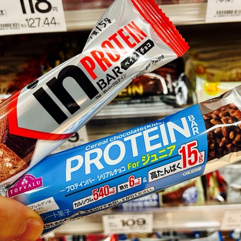 ムーンテッド（MoonTed）の日記より、スーパーの棚の前で手に持った2本のプロテインバー。森永のinバーと、トップバリューのプロテインバー