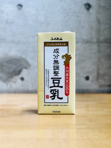 ムーンテッド（MoonTed）の日記より、ふくれんの「九州産大豆 成分無調整豆乳」の紙パック。コンクリート壁の前の木のテーブル