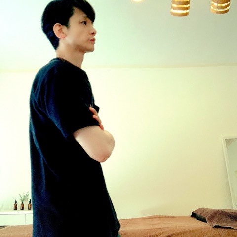 ムーンテッド（MoonTed）の日記より、施術台の横で腕を組んで立つムーンテッド（MoonTed）のセラピストAKI。黒いTシャツ姿の横顔