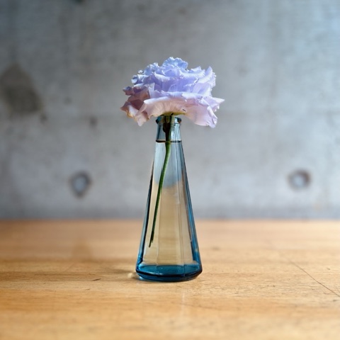 ムーンテッド（MoonTed）の日記より、あおのりの空き瓶を使った一輪挿しに、八重咲きの薄紫のトルコ桔梗。コンクリート壁の前の木のテーブル