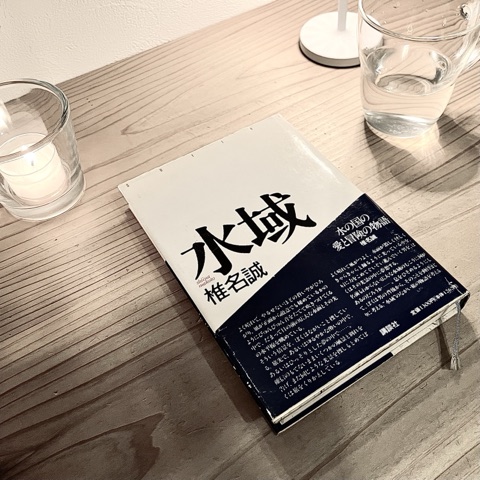ムーンテッド（MoonTed）の日記より、木のテーブルに置いた椎名誠の長編SF『水域』の単行本。そばに火をともしたキャンドルと水の入ったグラス